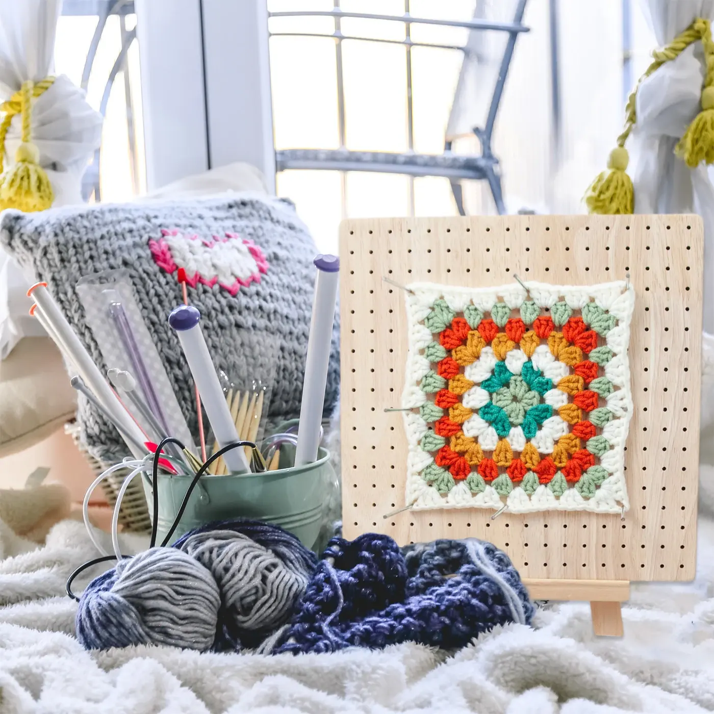 Square wooden blocking board holding a crochet square; yarn and knitting accessories shown for context are not included
